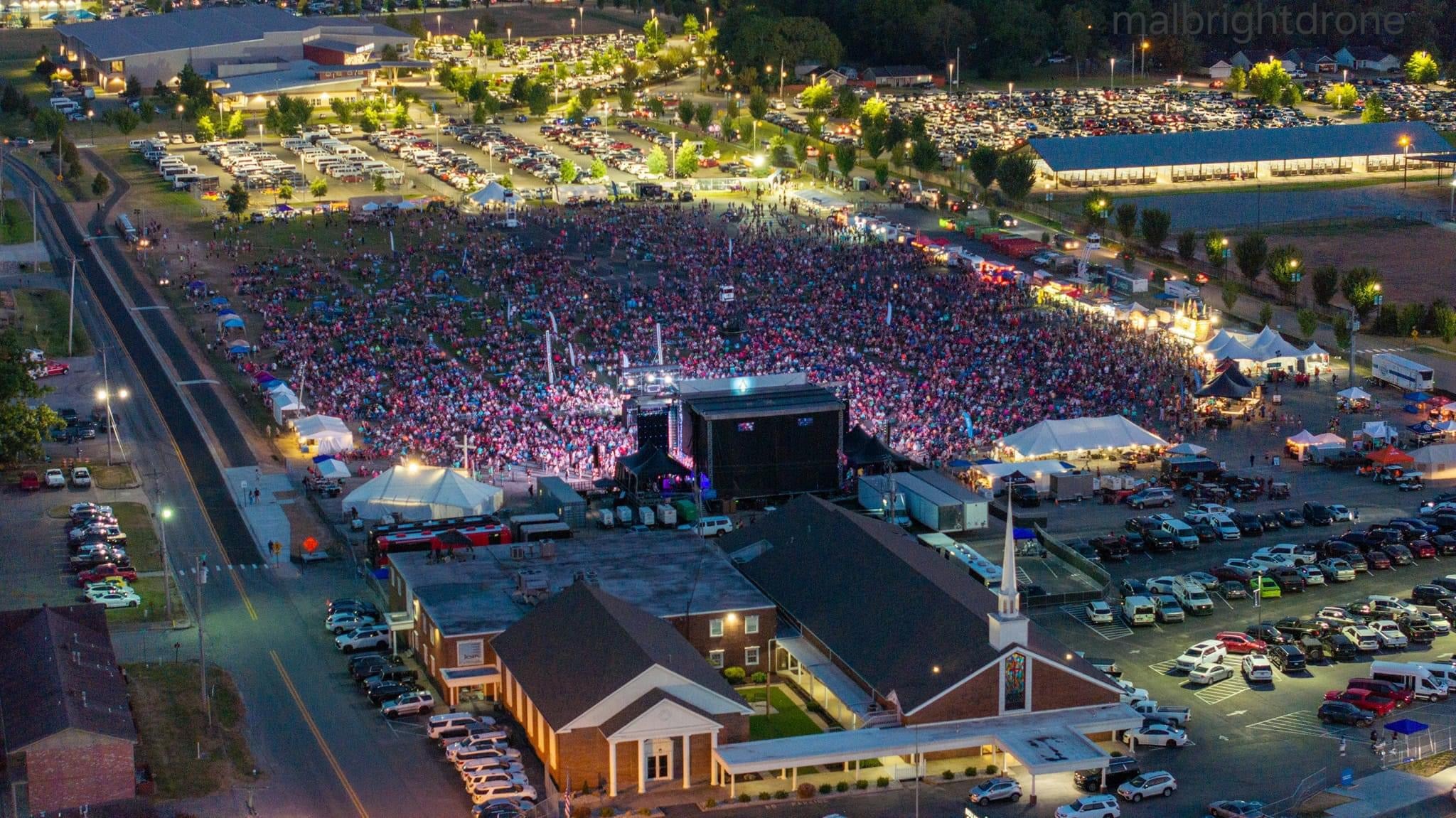 Aerial view of a massive outdoor concert at dusk, with thousands of people gathered in front of a brightly lit stage.