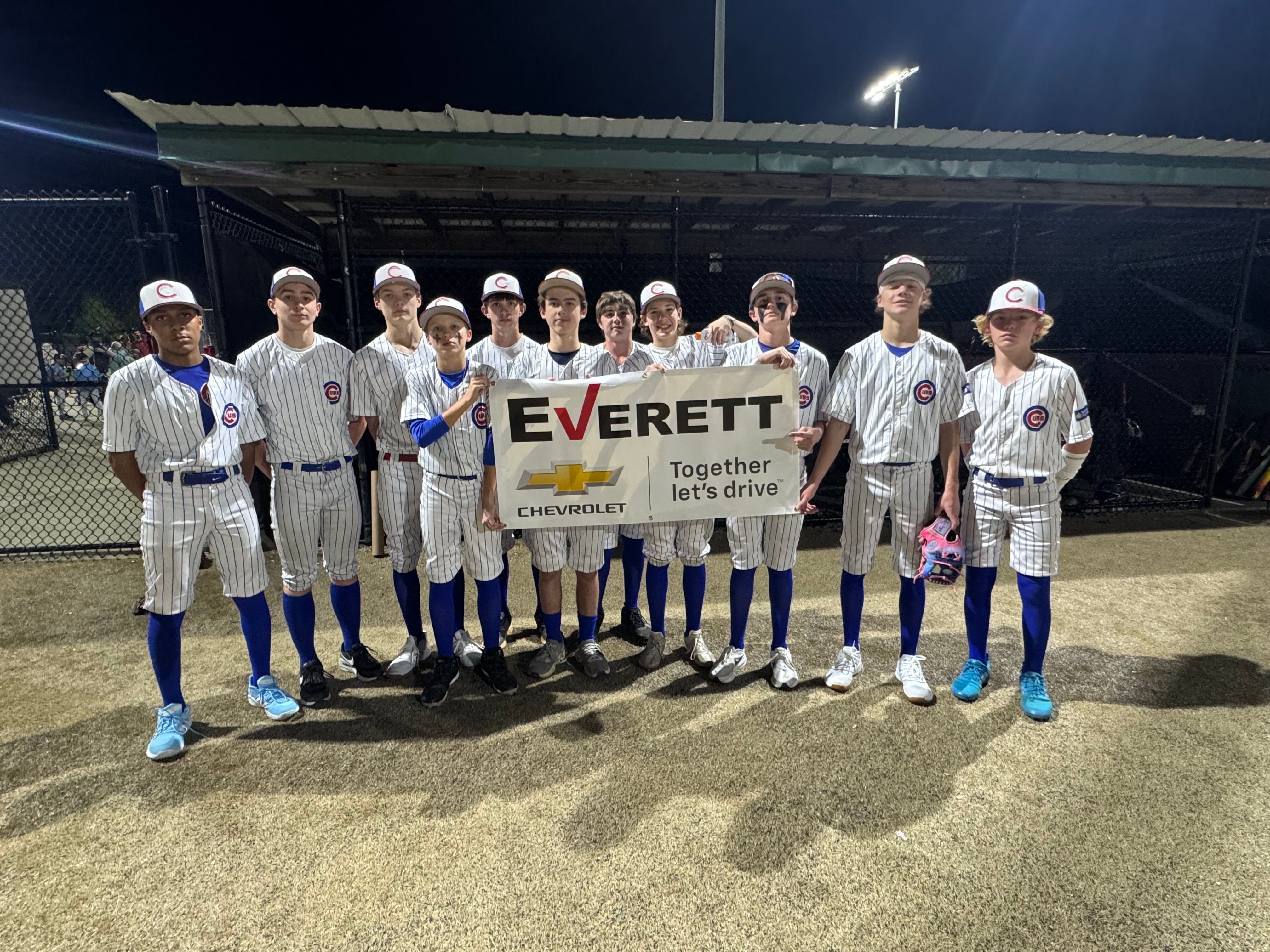 A youth baseball team in pinstripe uniforms and blue accessories holding a white Everett Chevrolet banner at night.