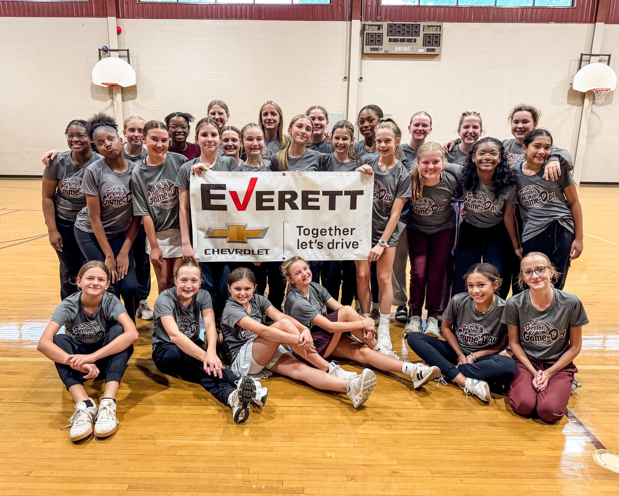 A group of smiling girls and young women in gray Benton Game-Day shirts pose in a gym with an Everett Chevrolet banner.