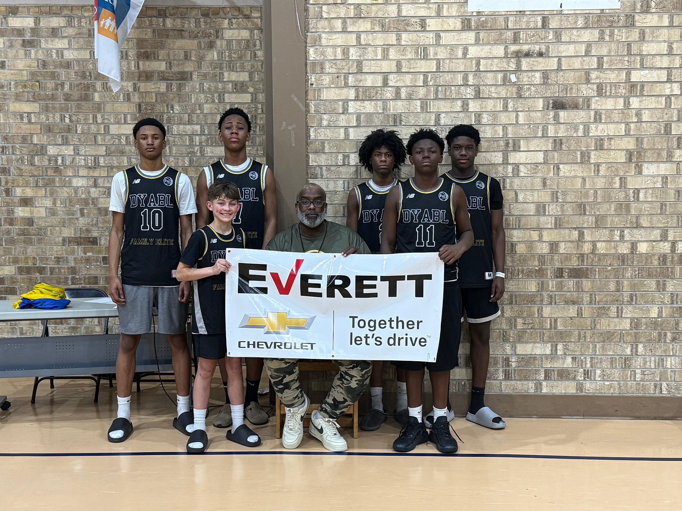 A basketball team of five young men and a coach pose with a long white banner for Everett Chevrolet.
